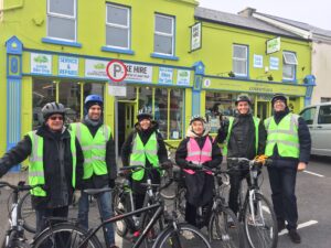A group of cheerful cyclists in high-visibility jackets ready for a ride, standing in front of a vibrant green building offering bike hire and repair services.