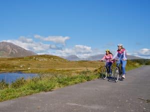 Two people cycle to work on a paved path by water, with mountains and blue sky behind them.