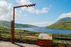 A scenic viewpoint overlooking a serene lake and rolling hills under a blue sky, marked by an artistic signpost indicating "killary harbour." an informational board is also present to educate visitors about the area.