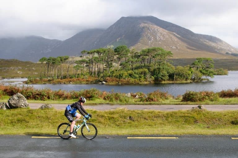 Cyclist rides scenic Road Bike Loops by a lake with a wooded island and mountains behind.