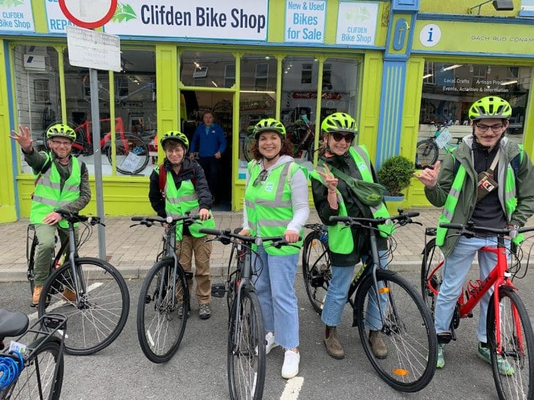 A group of five cyclists wearing high-visibility vests and helmets, ready for a ride, smiling outside the clifden bike shop.