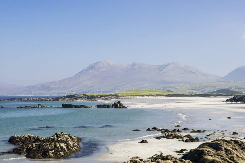 Sandy beach with rocks, calm Connemara sea, and distant mountains under a clear sky.