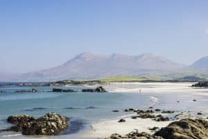 A serene beach with a lone individual walking along the shoreline, with scattered rocks in the shallow waters and majestic mountains rising in the background under a clear blue sky.