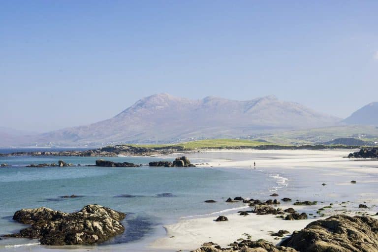 Sandy beach with rocks, calm Connemara sea, and distant mountains under a clear sky.