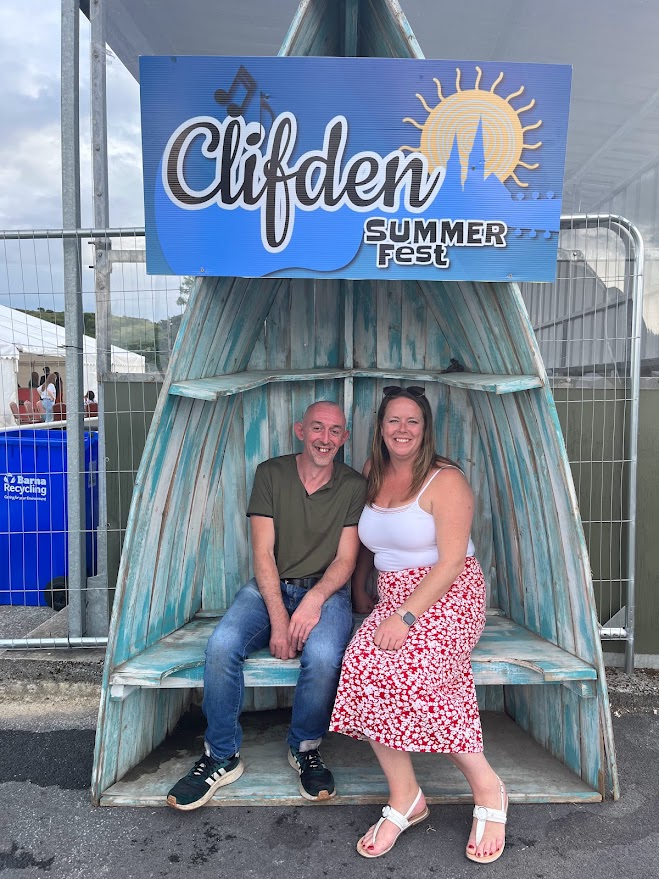 Two people smiling under a "Clifden Summer Fest 2025" sign in a wooden photo booth.