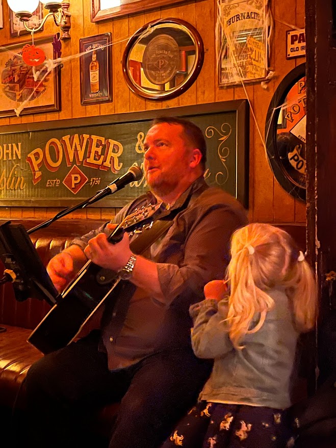 Man playing guitar at a pub during the Music and Session Trail Festival as a girl watches.