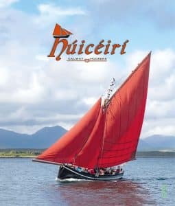 A traditional galway hooker sailboat with vibrant red sails on a calm sea, mountains in the background, under a cloudy sky.