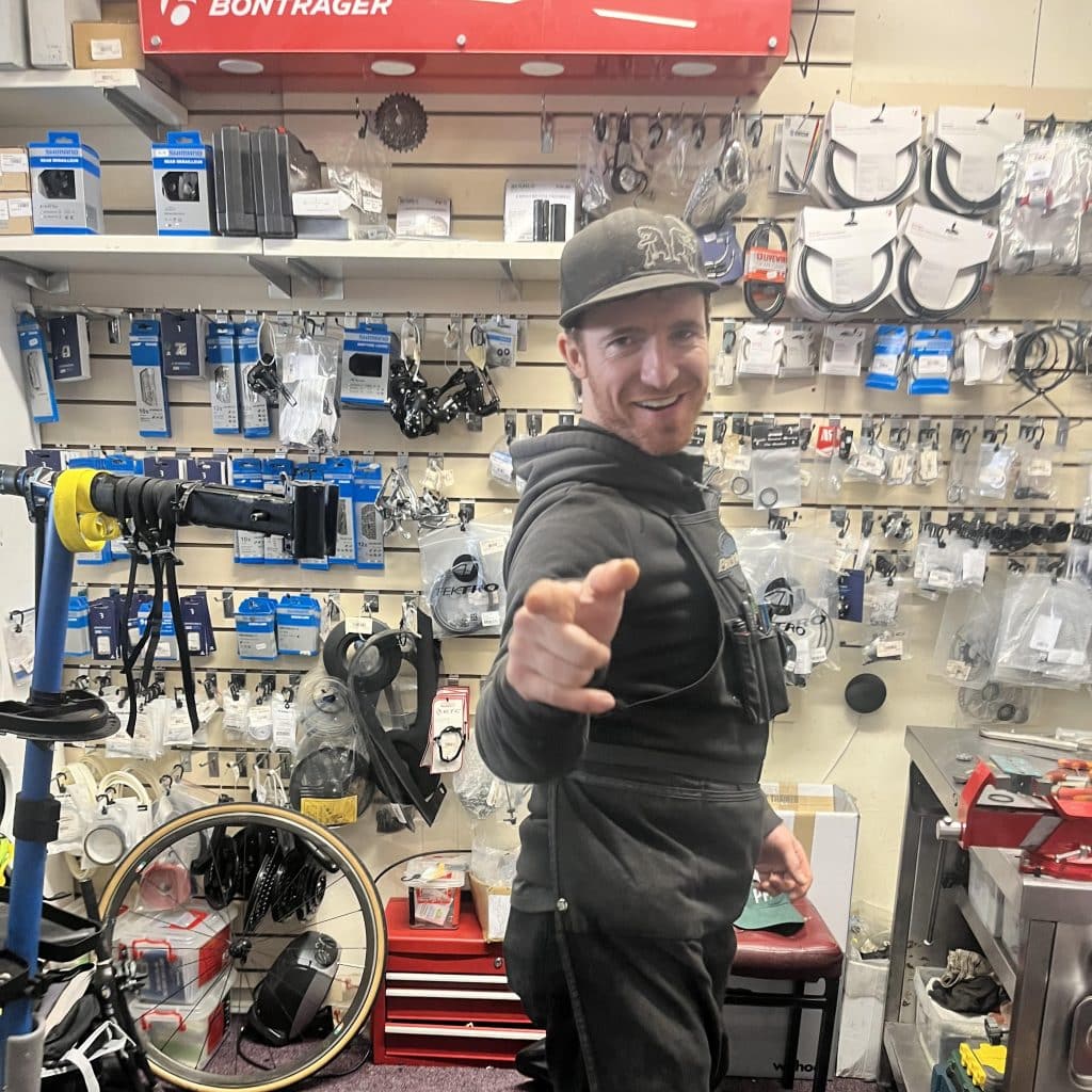 Stuart Connell, bicycle mechanic, smiling and pointing in a repair shop with tools behind him.