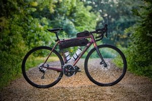 A red and black road bike with two Water Bottle Cages, Bontrager Elite - Black and a small bag attached to the frame is parked on a gravel path in a lush, green forest.