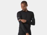 A person wearing a black cycling jersey with full-length zip stands against a plain background. They are adjusting their Bontrager Thermal Cycling Arm Warmers with a focused expression. The jersey has small logos on the sleeves and the arm warmers cover from wrist to bicep.
