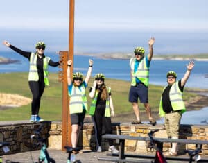 Five people in safety vests and helmets with Clifden Bike Hire bikes wave at a coastal viewpoint.