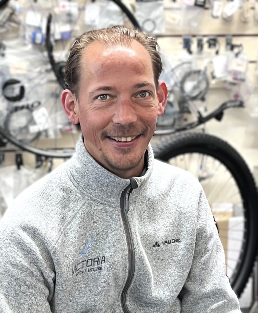 A smiling man in a light grey fleece sits by bicycle wheels at Clifden Bike Hire.