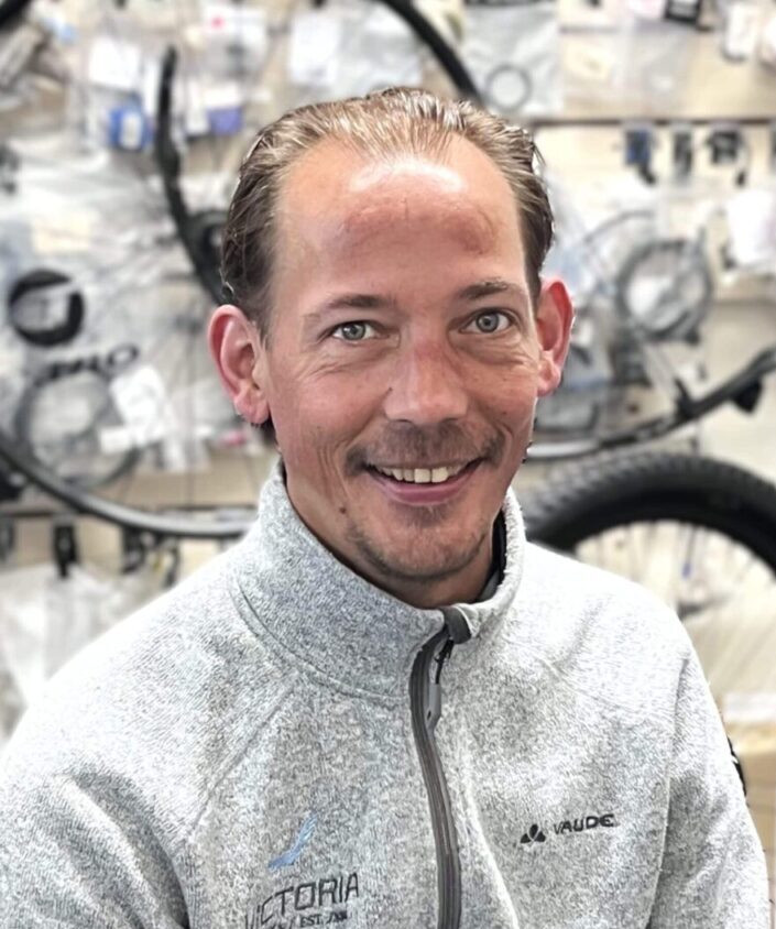 A smiling man in a light grey fleece sits by bicycle wheels at Clifden Bike Hire.