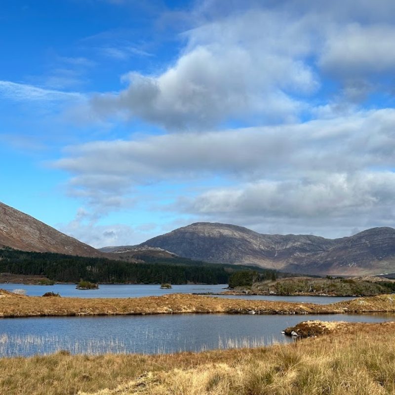 Serene lake nestled amidst rolling hills under a canvas of blue sky dotted with fluffy clouds.